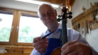 Der Geigenbaumeister Ralf Schumann hat eine Geige in der Hand. Er hat einen kleinen Holzstab in der Hand, um die Geige abzuklopfen. 