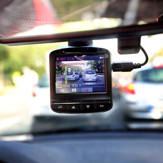 Eine Dashcam hängt vom Rückspiegel eines Autos herab, dahinter ist unscharf der Verkehr vor dem Auto zu erkennen. (Symbolbild): Dashcams können bei Unfällen wichtiges Beweismaterial liefern. Der Einsatz kann aber auch schnell illegal werden.