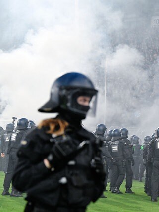 Ein Polizist steht beim Fußballspiel zwischen Waldhof Mannheim und dem 1. FC Kaiserslautern auf dem Platz. Im Hintergrund stehen zahlreiche weitere schwarz gekleidete Polizisten auf dem Rasen. Hinter ihnen steigt Rauch auf.