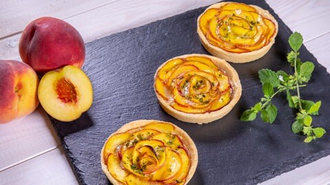Für Pfirsiche gibt es süße oder herzhafte Rezepte: Drei frisch gebackene Pfirsichtartelettes nebeneinander aufgereiht, ein angeschnittener Pfirsich und Minze daneben
