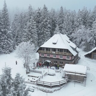 Gäste in einer Skibar genießen Après-Ski-Stimmung, lachen und stoßen miteinander an, während im Hintergrund die gemütliche Hüttengaststube zu sehen ist.