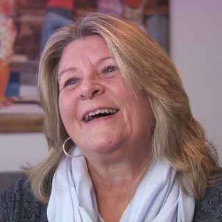 Silvia sitzt im Wohnzimmer. Sie hat blonde, schulterlange Haare und lächelt. 