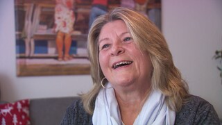 Silvia sitzt im Wohnzimmer. Sie hat blonde, schulterlange Haare und lächelt. 