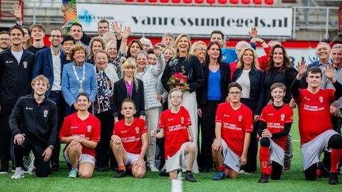 Königin Máxima steht auf dem Rasen eines Stadions inmitten von Menschen, vor ihr hocken Kinder in roten Trikots.