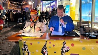 Zwei Blindenreporter sitzen an einem Tisch und sprechen in Mikrofone. Damit auch Menschen mit Sehbehinderung das närrische Treiben miterleben können, sind Blindenreporter beim Butzenlauf in Konstanz dabei.