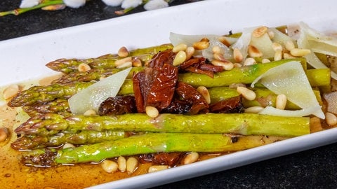 Eine weiße Platte mit grünem Spargel, auf dem Pinienkerne verstreut sind, zwischen den Spargelstangen liegen getrocknete Tomaten und oben drauf gehobelter, geschmolzener Parmesan
